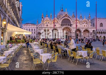 Le persone che siedono fuori dal caffè Lavena gustano un drink in Piazza San Marco in prima serata con la Basilica di San Marco sullo sfondo, Venezia, Italia Foto Stock