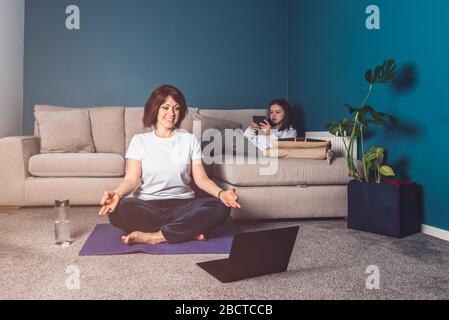 La donna sta facendo yoga online con il laptop durante l'isolamento di sé nel suo soggiorno, senza attrezzi di allenamento, suggerimenti per la meditazione per i principianti. Sua figlia sta leggendo. Famiglia con i bambini, rimanere a casa. Foto Stock