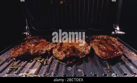 Deliziosa bistecca di carne succulenta cucinata alla griglia. Primi frittura di manzo su tostiera elettrica, rosmarino, pepe nero, sale. Movimento lento. Foto Stock