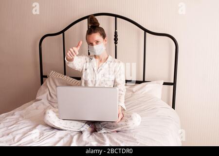 Ragazza giovane e felice che indossa la maschera medica tenendo il laptop in primo piano letto. Guardando la fotocamera. Assistenza sanitaria. Buongiorno. Studio a casa. Foto Stock