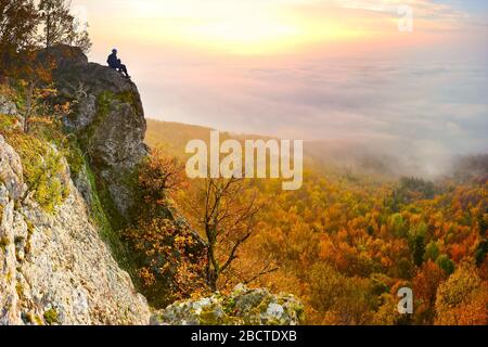 Alba con inversione sopra la profonda valle autunnale delle montagne con piccolo uomo in cima, che sta guardando questo bellissimo paesaggio Foto Stock