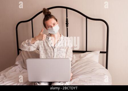 Donna felice che lavora a casa con un computer portatile a letto, con pigiama e maschera medica in camera. Guardando la fotocamera. Assistenza sanitaria. Buongiorno. Sociale dis Foto Stock