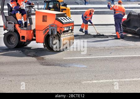 Gli addetti alla manutenzione stradale utilizzano un rullo vibrante, un bulldozer e una livella metallica per riparare la pavimentazione e compattare l'asfalto. Foto Stock