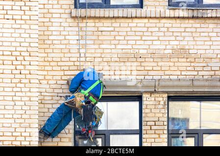 Costruttore scalatore appeso su funi di sicurezza sulla parete di un vecchio edificio con un set di diversi strumenti dietro. Foto Stock