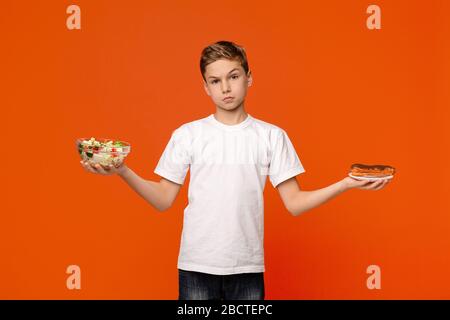 Triste ragazzo che sceglie tra insalata di verdure e cioccolato eclair Foto Stock