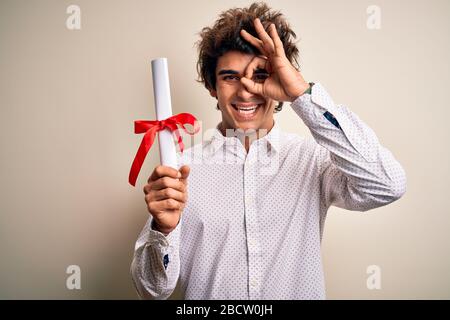 Giovane e bello studente che tiene il diploma in piedi su sfondo rosa isolato con felice viso sorridente fare ok segno con la mano sull'occhio guardando thru Foto Stock
