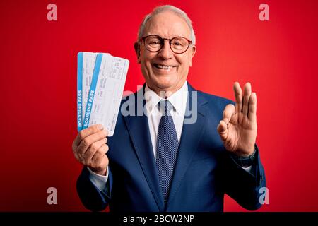 Uomo d'affari con i capelli grigi senior che tiene la carta d'imbarco dell'aeroplano sopra lo sfondo rosso che fa il segno giusto con le dita, simbolo eccellente Foto Stock