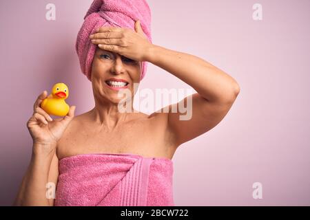 Donna di media età che indossa un asciugamano da bagno rosa dalla cura del corpo di bellezza che tiene l'anatra gialla di gomma stressata con la mano sulla testa, scioccata con vergogna e sorpresa Foto Stock