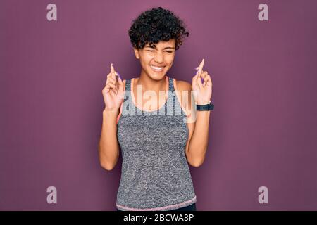 Giovane bella afroamericana afro sportswoman fare esercizio con l'abbigliamento sportivo gesturing dito attraversato sorridente con speranza e gli occhi chiusi. Fortuna a Foto Stock