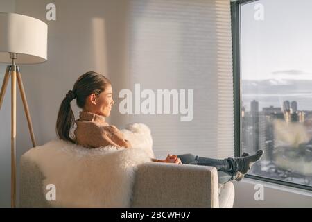 Rimanere a casa donna asiatica rilassante pensiva guardando fuori la finestra seduta in soggiorno divano godere del tempo libero per pensare e pratica la consapevolezza, al chiuso durante lo scoppio del coronavirus. Foto Stock