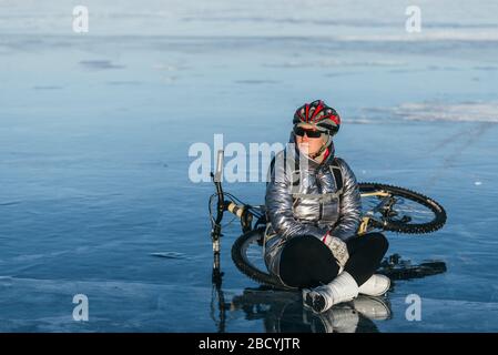 Donna seduto vicino alla sua bici sul ghiaccio. La ragazza ciclista si fermò a riposare. Si siede al volante e gode della splendida vista del tramonto. Foto Stock