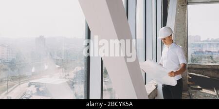 Uomo d'affari caucasico che tiene un piano di costruzione e lo analizza mentre posa vicino ad una grande finestra con un casco Foto Stock