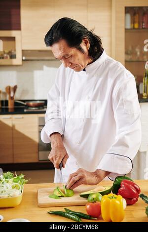 Serio uomo asiatico di mezza età che taglia avocado biologico fresco quando fa la cena a casa Foto Stock