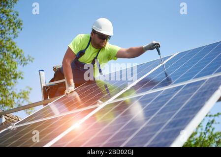 Tecnico che collega il pannello fotovoltaico solare alla piattaforma metallica utilizzando un cacciavite in piedi sulla scala su sfondo blu brillante dello spazio di copia cielo. Concetto di installazione del sistema a pannelli solari autonomi. Foto Stock