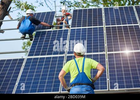 Vista posteriore del tecnico di ingegneria in piedi davanti al non finito alto impianto fotovoltaico pannello solare esterno con due lavoratori su piattaforma alta. Foto Stock