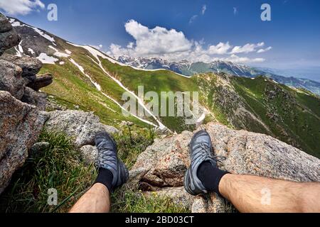 Gambe di uomo in scarpe di tracking e la vista delle montagne innevate con sfondo con cielo nuvoloso Foto Stock