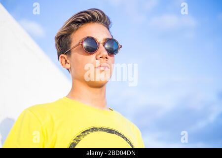 Bel ragazzo uomo bello con capelli biondi e occhiali da sole neri stand e posa con camicia gialla e cielo blu sfondo - caucasian Guy estate ritratto Foto Stock