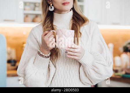 Foto di scorta tagliata di una giovane donna elegante in maglione accogliente e braccialetti tenendo una tazza rosa con strass nelle sue mani curate nel Foto Stock