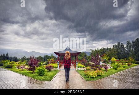 Woman in Red controllato shirt e hat andando alla Pagoda giapponese giardino alla pioggia cielo nuvoloso Foto Stock