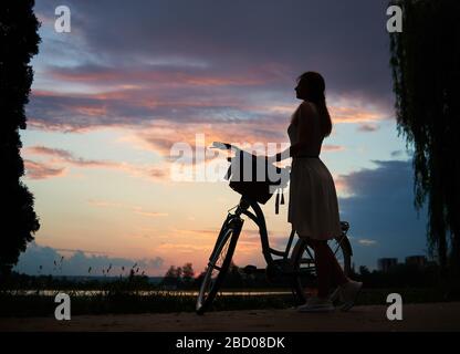 Donna in un abito leggero con bici retrò sotto il cielo al tramonto. Godendo di una splendida vista del cielo serale con nuvole colorate in estate. Il concetto di libertà e relax Foto Stock