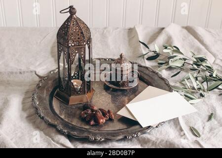 Biglietto d'auguri Ramadan Kareem, scena mockup invito. Piatto di bronzo con frutta datteri, tazza di tè, rami d'oliva e lanterna marocchina luminosa su lino Foto Stock