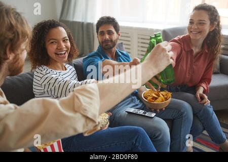 Ritratto di un gruppo multietnico di amici che attillano le bottiglie di birra mentre guardano la TV insieme seduti su un comodo divano a casa Foto Stock