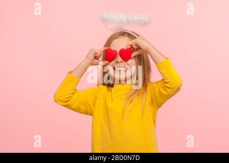 Ritratto di bella ragazza felice angelico con alone sopra la testa che copre gli occhi con i cuori giocattolo e sorridente sinceramente, metafora di sguardo pieno di amore. ind Foto Stock