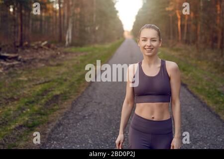 Sorridente atletica in forma giovane donna che si allinea su una corsia asfaltata attraverso le foreste retroilluminata dal caldo bagliore del sole in uno stile di vita sano e attivo concep Foto Stock