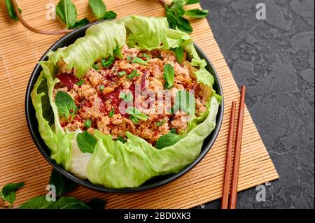 Laab Gai - insalata di pollo sminuzzato tailandese in ciotola nera su sfondo di ardesia scuro. Larb è piatto della cucina tailandese con carne tritata, salsa di pesce, succo di lime, m Foto Stock