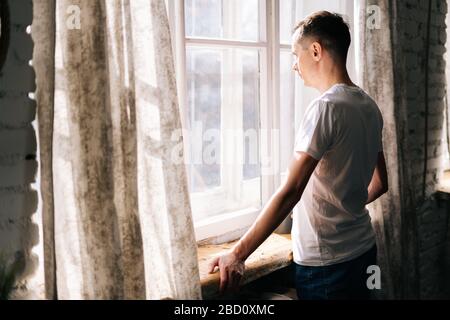 Il giovane uomo premuroso si alza accanto alla finestra durante il giorno di sole. Foto Stock