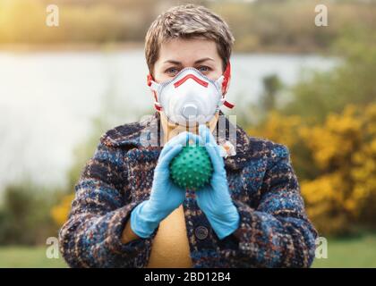 Morbido fuoco di ritratto di una donna che indossa una maschera facciale bianca e rossa, guanti blu, cappotto blu, giallo top e tenendo coronavirus nelle sue mani Foto Stock