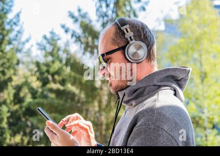 Uomo adulto bearded in felpa con cappuccio con cuffie wireless che prendono il selfie con smartphone nel parco all'aperto, testa e spalle Foto Stock