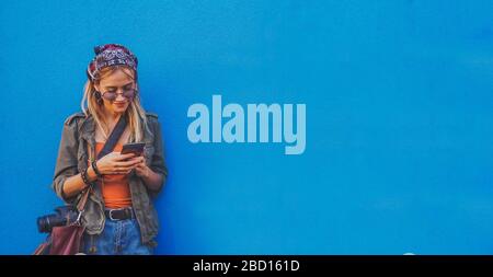 Giovane ragazza in piedi sullo sfondo blu con uno smartphone - UN hipster elegante ragazza sorridente e di controllo dei social media Foto Stock