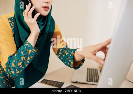 Immagine ritagliata della signora di affari musulmana in vestiti tradizionali che puntano allo schermo del calcolatore e che parlano sul telefono con il collega Foto Stock