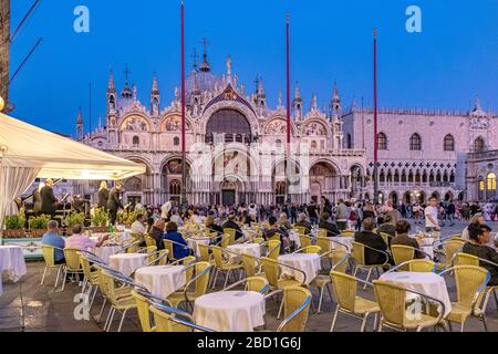 Le persone che siedono fuori dal caffè Lavena gustano un drink in Piazza San Marco in prima serata con la Basilica di San Marco sullo sfondo, Venezia, Italia Foto Stock