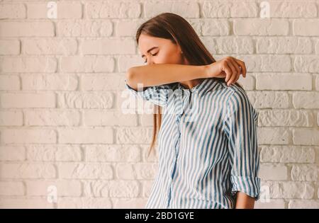 Giovane donna malata che copre il naso e la bocca con il braccio mentre starnutisce a casa. Paziente infetto malato della ragazza tosse nel suo gomito. Coronavirus COVID-19 sy Foto Stock