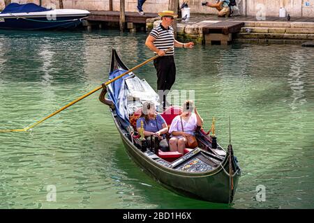 Una gondola in uniforme gondolieri gusterai la sua gondola lungo un canale con due turisti femminili a bordo, Venezia, Italia Foto Stock