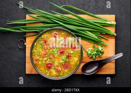 Zuppa di piselli secca con salsicce di carne, patate, cipolle verdi e sesamo. Zuppa di piselli gialla in una ciotola nera su sfondo di ardesia scura. Foto Stock