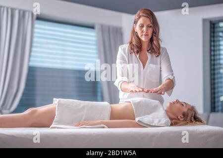 Giovane donna rilassata coperta da asciugamani con guarigione reiki con un terapista concentrato con occhi chiusi. Foto Stock