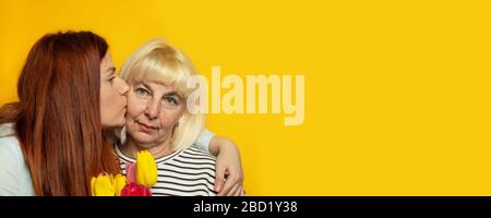 Adulto bella mamma dolce che tiene freschi tulipani fiori e sta ricevendo un bacio da sua figlia giovane con spazio copia su sfondo muro giallo Foto Stock