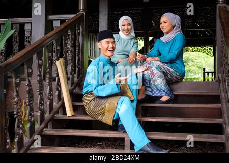 Un gruppo di tre musulmani malesi in costume tradizionale stanno avendo una conversazione felice durante la celebrazione di Aidilfitri sulla terrazza di legno tradizionale Foto Stock