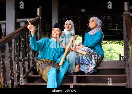 Un gruppo di tre musulmani malesi in costume tradizionale stanno avendo una conversazione felice durante la celebrazione di Aidilfitri sulla terrazza di legno tradizionale Foto Stock