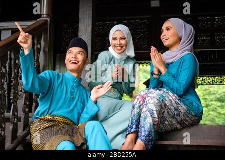 Un gruppo di tre musulmani malesi in costume tradizionale stanno avendo una conversazione felice durante la celebrazione di Aidilfitri sulla terrazza di legno tradizionale Foto Stock