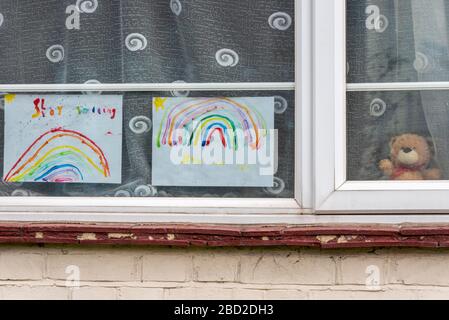 L'arte dell'arcobaleno del bambino e l'orso del teddy nella finestra della casa durante il periodo di blocco di pandemia di Coronavirus di COVID-19. Caccia all'orso Teddy, sentiero dell'arcobaleno Foto Stock