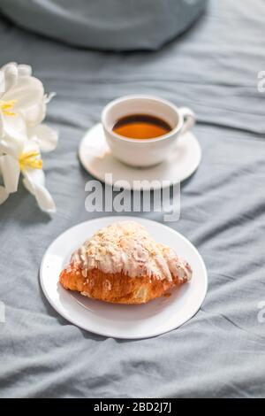 Croissant fresco su un piatto di vetro bianco e una tazza di caffè sul letto. Colazione a letto. Bouquet di tulipani bianchi. Foto verticale Foto Stock