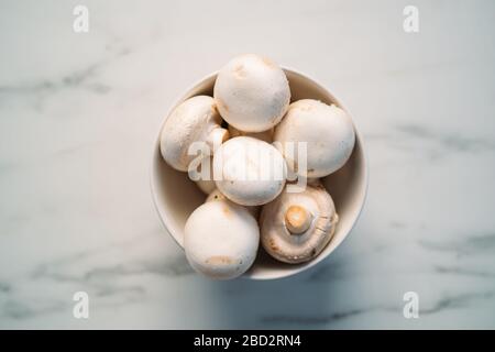 Set di funghi ​​champignons freschi interi e tagliati in una ciotola isolata su fondo di pietra bianca. Vista dall'alto Foto Stock