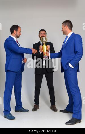 Uomini d'affari con facce felici in abiti formali tengono il premio d'oro su sfondo grigio. I colleghi tengono la tazza come simbolo della vittoria. I leader celebrano la competizione aziendale vincente. Successo nel concetto di business Foto Stock