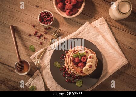 Pancake con lamponi, fette di banana, semi di melograno e miele su tavola d'annata in legno. Foto Stock