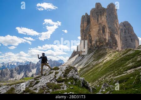 Vista grandangolare di un paesaggio montano estivo, con un escursionista che prende un selfy in primo piano e le tre cime di Lavaredo sullo sfondo Foto Stock