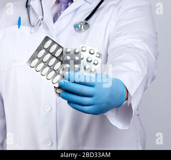 il medico terapista è vestito con un cappotto uniforme bianco e guanti sterili blu è in piedi e in possesso di una pila di pillole in confezioni blister, bianco backgroun Foto Stock